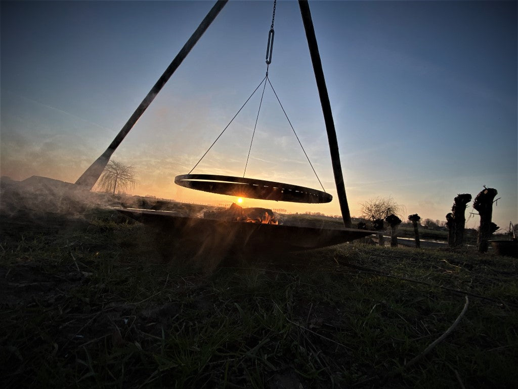 Een ondergaande zon met op de voorgrond een houten driepoot van Basale. De zon schijnt precies tussen de vuurschaal en het grillrooster door