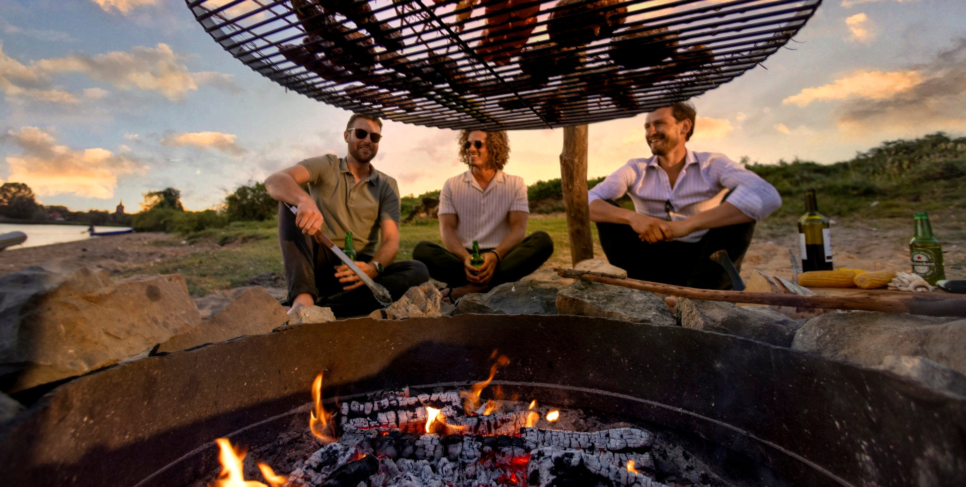 Een foto van 3 vrienden die aan het barbecuen zijn boven een echt vuur. Boven het vuur hangt een grillrooster van Basale aan een zelfgemaakte driepoot barbecue met de DIY tripod